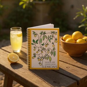 Könnte beinhalten: Ein Spiralnotizbuch mit gelbem Rand und Zitronenillustration. Auf dem geöffneten Notizbuch steht "WHEN LIFE GIVES YOU...LEMONS". Ein Glas Limonade, eine Zitronenhälfte und eine Schüssel mit Zitronen sind ebenfalls zu sehen.