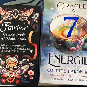 May include: Two oracle card decks. The first, "Fairies Oracle Deck and Guidebook," features a black cover with a fairy illustration and text. The second, "Oracle of the 7 Energies," has a colorful bowl illustration and text.