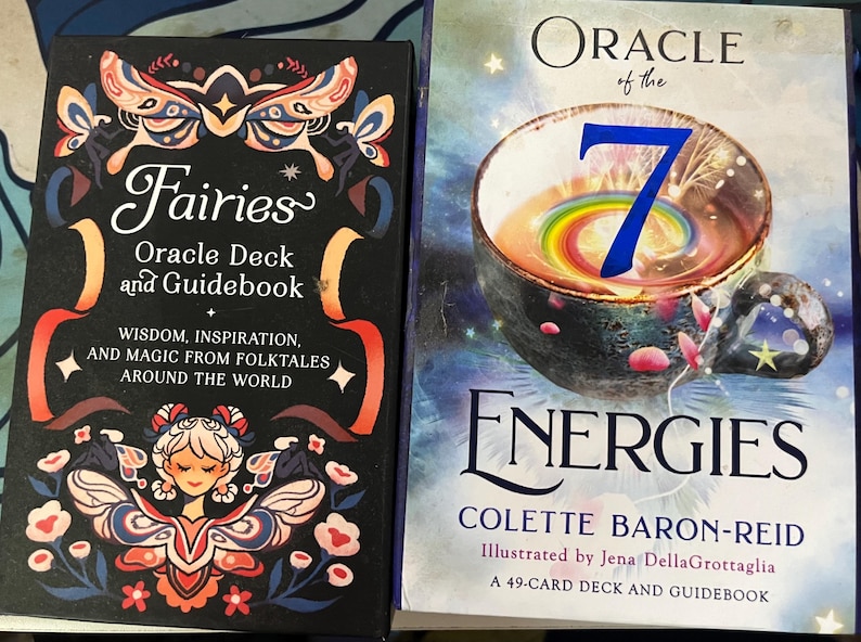 May include: Two oracle card decks. The first, "Fairies Oracle Deck and Guidebook," features a black cover with a fairy illustration and text. The second, "Oracle of the 7 Energies," has a colorful bowl illustration and text.