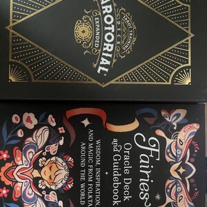 May include: Two tarot card boxes. The top box is black with gold geometric designs and the text "TAROTORIAL EXPANDED" and "TAROT TRAINING DECK". The bottom box is black with a colorful fairy illustration and the text "Fairies Oracle Deck and Guidebook".