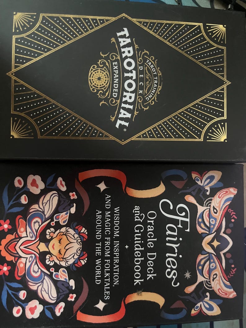 May include: Two tarot card boxes. The top box is black with gold geometric designs and the text "TAROTORIAL EXPANDED" and "TAROT TRAINING DECK". The bottom box is black with a colorful fairy illustration and the text "Fairies Oracle Deck and Guidebook".