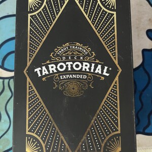 May include: A black tarot card deck box with gold geometric designs and the text "Tarot Training Deck Tarutorial Expanded". The box has a diamond-shaped design in the center with radiating lines and dotted patterns.