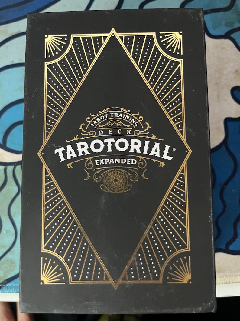 May include: A black tarot card deck box with gold geometric designs and the text "Tarot Training Deck Tarutorial Expanded". The box has a diamond-shaped design in the center with radiating lines and dotted patterns.