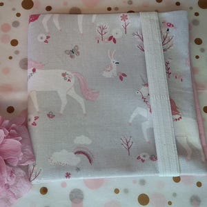 Könnte beinhalten: Ein hellgraues Stoffobjekt mit einem Einhorn-Muster, das rosa Mähnen und Schwänze, Schmetterlinge und Regenbogen zeigt. Ein weißes Gummiband sichert das Objekt. Zwei rosa Blumen flankieren das Objekt, vor einem gepunkteten Hintergrund.
