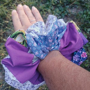 以下が含まれることがあります： さまざまな花柄と無地のパターンが施された、カラフルなシュシュのコレクション。シュシュは、紫、青、白の色合いで、花柄プリントと無地の生地が特徴です。これらのヘアアクセサリーは、スタイルのアクセントを加えるようにデザインされています。