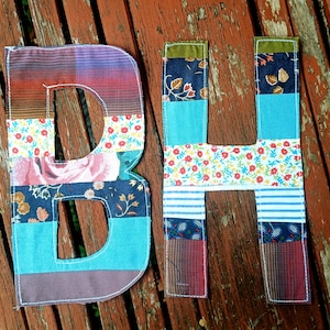 Op de afbeelding: Twee stoffen letters, B en H, gemaakt van diverse patchwork textielstroken. De letters bevatten een mix van bloemen-, streep- en effen patronen in tinten blauw, rood, roze en bruin, met zichtbare witte stiksels langs de randen.