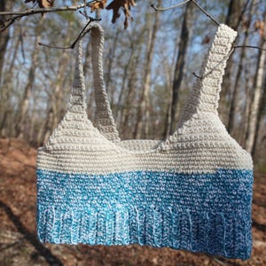 Op de afbeelding: Een gehaakt topje met wit en blauw garen. De top heeft een wit bovenste gedeelte en een blauw onderste gedeelte. De bandjes zijn wit.