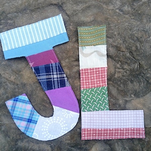 May include: A patchwork fabric letter "J" and "L" set against a textured, gray stone surface. The letters are composed of various fabric patterns, including stripes, plaid, and floral designs in shades of blue, purple, green, and red.