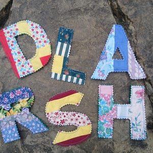 May include: Fabric letters spelling "DORASH" are displayed. Each letter is crafted from patchwork fabric, featuring floral and geometric designs, and finished with scalloped edges. The letters are arranged on a textured, grey stone surface.