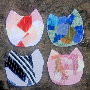 May include: Four fabric patches shaped like cat heads, each featuring a unique patchwork design. One patch uses red, pink, and white fabric scraps, another uses blue and green tones, one features black and white stripes, and the last uses pink plaid.