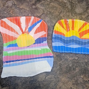 May include: Two fabric patches featuring a sunrise design. The larger patch displays a yellow sun with orange and white rays above horizontal bands of green, pink, blue, and white. The smaller patch shows a yellow sun with orange and yellow rays above wavy blue stripes.