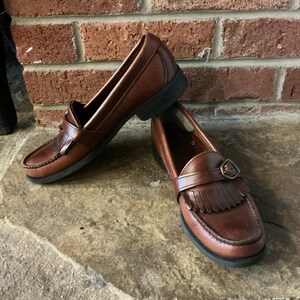 Può includere: Un paio di mocassini in pelle marrone dal design classico. Le scarpe presentano uno stile slip-on con cinturino e fibbia, e un dettaglio con frange. Le suole sono nere. Le scarpe sono su uno sfondo di mattoni.