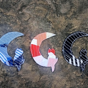 Pode incluir: Três emblemas em forma de lua crescente e gato feitos de retalhos de tecido. O emblema da esquerda apresenta padrões de riscas azuis e brancas. O emblema central exibe segmentos laranja, vermelhos e brancos. O da direita é preto com riscas.