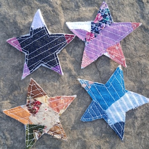 May include: Four fabric stars featuring patchwork designs. One star is black with white stripes, one is purple and floral, one is multi-coloured with floral and geometric patterns, and one is blue with various striped and solid denim-like textures.