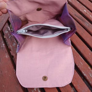 Puede incluir: Bolso de tela hecho a mano, abierto para mostrar el interior rosa claro y un bolsillo con cremallera. El exterior presenta un estampado de cuadros morados y rosas. Cierre de botón a presión de latón visible. El bolso está abierto.