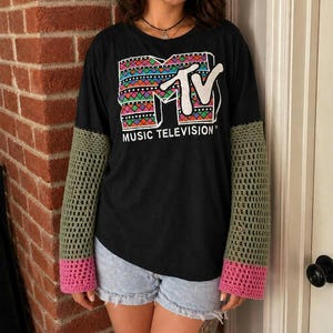 May include: Black long-sleeve shirt with a colorful MTV logo and the words "MUSIC TELEVISION". The sleeves are olive green crochet with pink cuffs. The shirt is paired with light blue denim shorts.