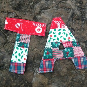 Op de afbeelding: Rode en groene stoffen letters die "TA" spellen met een kerstthema. De "T" is rood met witte stippen en een wit kerstornamentontwerp. De "A" is rood met een wit cadeauontwerp en hulstbladeren.
