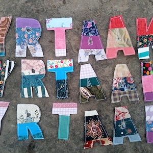 Könnte beinhalten: Eine Ansammlung von Stoffbuchstaben, die "YORKTA" buchstabieren, auf einer strukturierten Oberfläche angeordnet. Die Buchstaben sind Patchwork, mit verschiedenen floralen und gemusterten Stoffen in Rosa, Blau, Lila und Grün. Die Buchstaben sind handgefertigt.