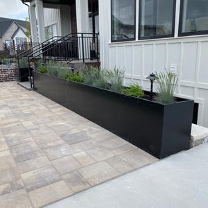 May include: A long, rectangular black planter box filled with green and gray plants. The planter is positioned in front of a white house with black trim and windows. The ground is paved with light-colored rectangular stones.