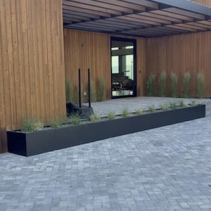 May include: Exterior view of a modern building with a long, black rectangular planter filled with greenery. The building features wooden siding and a dark-colored pergola over the entrance. The ground is paved with gray bricks.