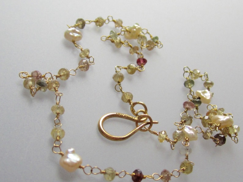 May include: Delicate gold-toned necklace featuring a series of small, colorful gemstones and irregular-shaped pearls. The necklace is strung together with fine gold wire and has a gold clasp. The gemstones are in shades of yellow, pink, and brown.