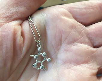 May include: A silver necklace with a small, detailed pendant shaped like a caffeine molecule. The pendant has small clear stones for the atoms.