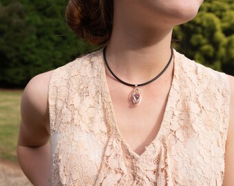 May include: A woman wearing a beige lace top with a black choker necklace and a silver pendant featuring a clear crystal.