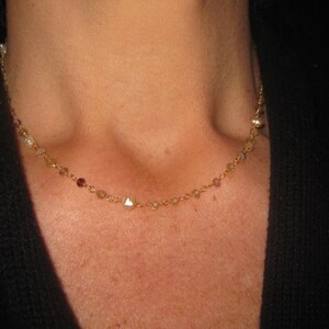 May include: Delicate gold necklace with small, colorful beads and pearls. The necklace is draped around a person's neck, showcasing its fine details and elegant design. The necklace is a beautiful piece of jewelry.
