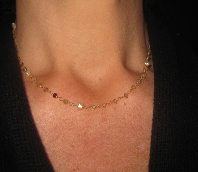 May include: Delicate gold necklace with small, colorful beads and pearls. The necklace is draped around a person's neck, showcasing its fine details and elegant design. The necklace is a beautiful piece of jewelry.