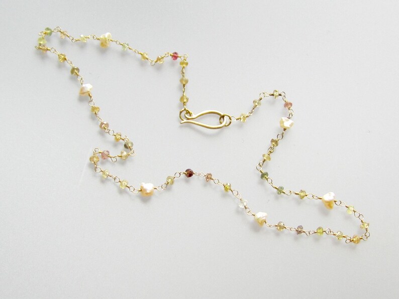 May include: Delicate gold necklace featuring small, colorful gemstones and irregular pearls. The necklace has a delicate chain and a gold-colored clasp. The gemstones are in shades of yellow, green, and pink, creating a subtle, elegant design.