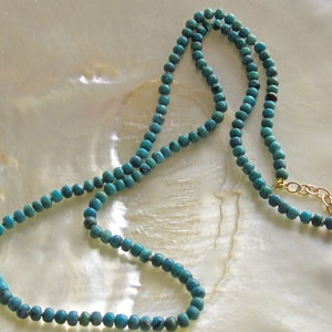 Op de afbeelding: Een turquoise blauwe kralenketting met een gouden kettingsluiting. De ketting ligt op een wit parelmoer oppervlak.