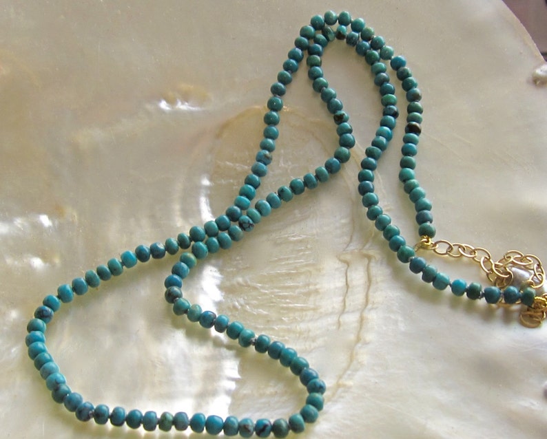 Op de afbeelding: Een turquoise blauwe kralenketting met een gouden kettingsluiting. De ketting ligt op een wit parelmoer oppervlak.