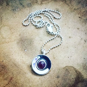May include: A silver necklace with a round pendant featuring a deep purple gemstone set in a silver circle.