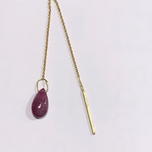 Może przedstawiać: Złoty kolczyk z łańcuszkiem, z wiszącym z niego wisiorem w kształcie łzy z rubinu i długim, cienkim prętem ze złota.