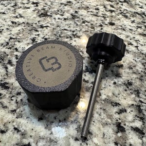 May include: A black knob with a threaded rod next to a cylindrical object with the text "CREATIVE BEAM STUDIO" and a logo. The cylindrical object is black with a tan top.