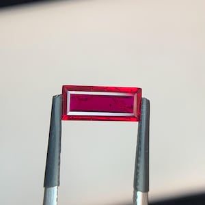 May include: A vibrant, deep red gemstone cut into a rectangular baguette shape. The stone is held by a pair of dark grey metal tweezers, showcasing its transparent quality and sharp, clean edges. The surface of the gem reflects light clearly.