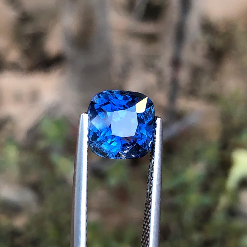 May include: A square-cut, deep blue sapphire gemstone held by metal tweezers. The sapphire's facets catch the light, creating a sparkling effect. The background is blurred, focusing attention on the gem's colour and clarity.