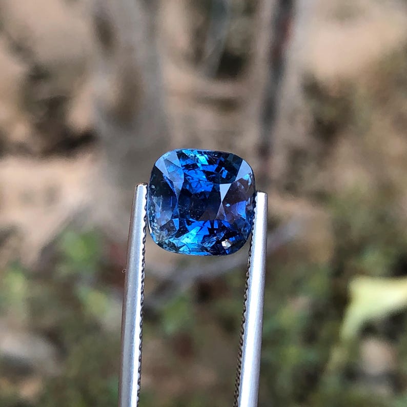 May include: A square-cut, deep blue gemstone held by silver tweezers. The gem's facets catch the light, displaying a rich blue colour. The blurred background accentuates the gemstone's clarity and colour. This image showcases a precious stone.