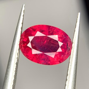 May include: A vibrant, oval-cut ruby gemstone held by silver tweezers. The ruby displays a deep red hue with a star-like pattern in the center. The gemstone is set against a neutral background, highlighting its color and clarity.