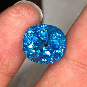 May include: A brilliant, cushion-cut gemstone with a vibrant, light blue hue. The facets of the gem reflect light, creating a dazzling display of color and sparkle. The gem is held between two fingers, showcasing its size and clarity.