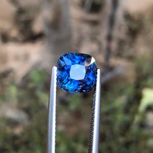 May include: A square-cut, deep blue gemstone is held by silver tweezers. The gem's facets catch the light, creating a sparkling effect. The background is out of focus, emphasizing the gem's colour and clarity. This image presents a precious stone.