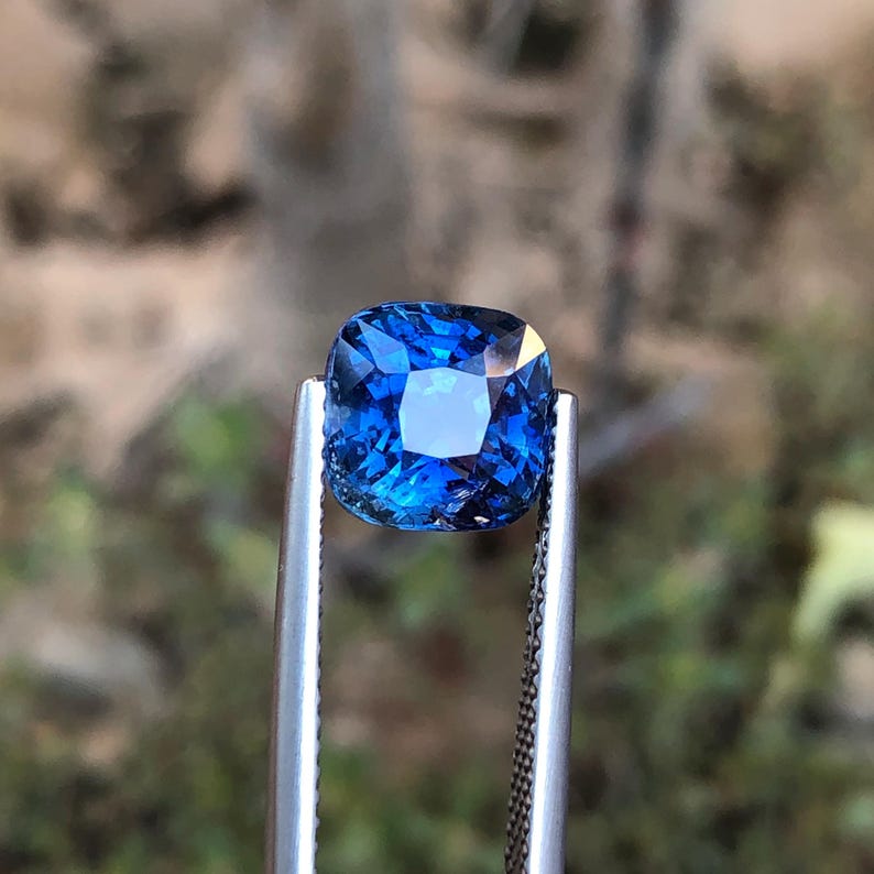 May include: A square-cut, deep blue gemstone is held by silver tweezers. The gem's facets catch the light, creating a sparkling effect. The background is out of focus, emphasizing the gem's colour and clarity. This image presents a precious stone.