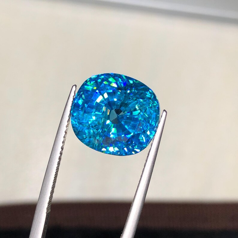 May include: A vibrant, cushion-cut gemstone held by silver tweezers. The gem is a brilliant shade of turquoise, with facets that catch and reflect light, creating a sparkling effect. The gem's shape is rounded with a flat top and bottom.