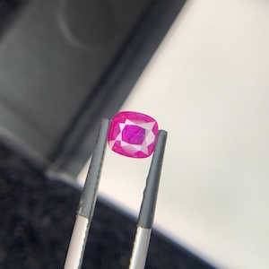 Może przedstawiać: Rubin o szlifie poduszkowym, trzymany pęsetą. Rubin ma głęboki, różowo-czerwony odcień z wewnętrznymi fasetami odbijającymi światło. Kamień szlachetny jest umieszczony na neutralnym tle, podkreślając jego kolor i przejrzystość.