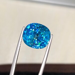 May include: A brilliant, cushion-cut gemstone held by tweezers. The gem is a vibrant, sparkling blue with multiple facets that catch the light. The gem's shape is a rounded square, and it appears to be a high-quality, precious stone.
