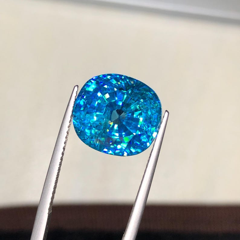 May include: A brilliant, cushion-cut gemstone held by tweezers. The gem is a vibrant, sparkling blue with multiple facets that catch the light. The gem's shape is a rounded square, and it appears to be a high-quality, precious stone.