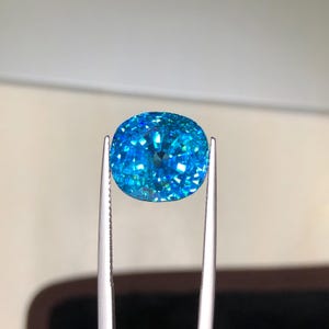 May include: A vibrant, cushion-cut gemstone held by silver tweezers. The gem is a brilliant shade of blue, with facets that reflect light, creating a sparkling effect. The background is a soft, neutral tone, highlighting the gem's color and clarity.