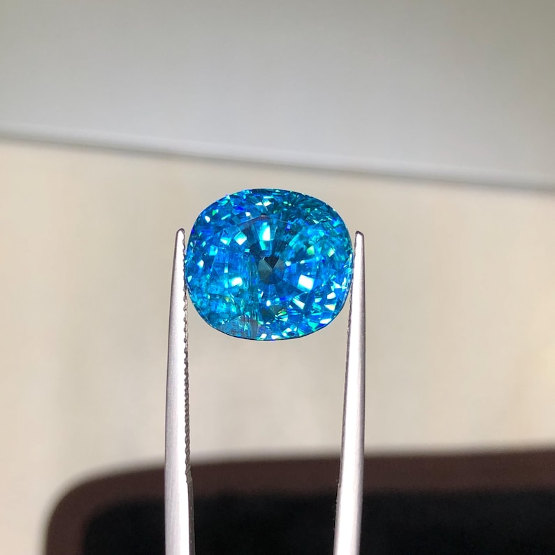 May include: A vibrant, cushion-cut gemstone held by silver tweezers. The gem is a brilliant shade of blue, with facets that reflect light, creating a sparkling effect. The background is a soft, neutral tone, highlighting the gem's color and clarity.