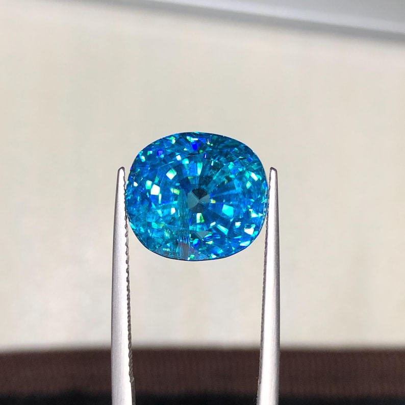 May include: A brilliant, oval-cut gemstone with a vibrant, light blue hue. The gem is held by silver tweezers, showcasing its facets and internal brilliance. The stone's color shifts with light, creating a captivating display of blues and greens.