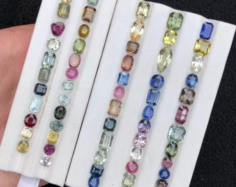 May include: A collection of loose faceted gemstones arranged in five vertical rows on a white display board. The stones feature various shapes including round, oval, square, and emerald cuts, showcasing a spectrum of colors like blue, yellow, green, pink, and clear.
